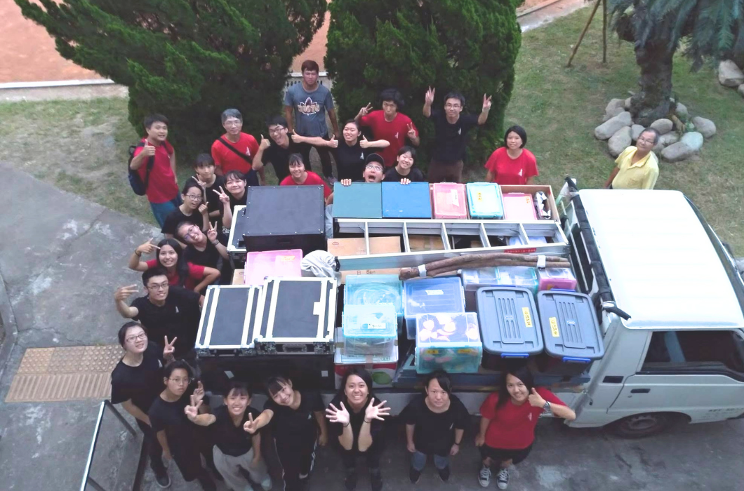 劇團工作人員與滿載道具的小貨車合影。俯瞰視角拍攝，貨車後車斗整齊堆滿了大型黑色音響道具箱與各式收納盒，約二十名身穿紅、黑團服的年輕團員圍繞在車旁開心比讚揮手。