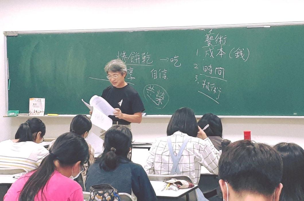 室內排練或講座現場。一位灰白頭髮、戴眼鏡的男性講師正站在黑板前翻閱資料，黑板上寫著「烤餅乾」、「藝術：1.成本（錢） 2.時間 3.技術」等字樣，台下有多位學員正專注聽講。