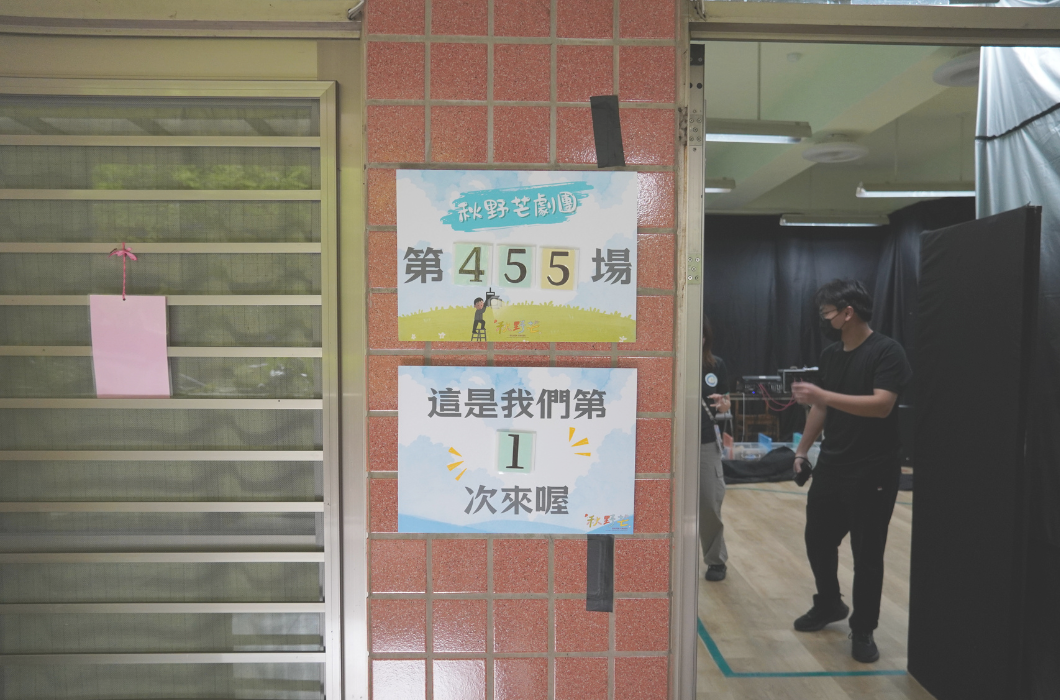 教室門口的活動告示牌。紅磚柱上貼著兩張海報，上方寫著「秋野芒劇團 第455場」，下方寫著「這是我們第1次來喔」，門內可見工作人員正在準備舞台。