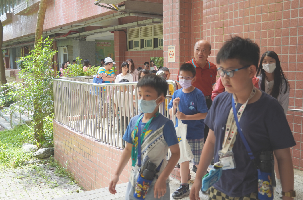 學童排隊入場的畫面。多位戴著口罩、掛著水壺的國小學生在老師與志工的引導下，正順著校舍走廊與無障礙坡道依序前進。  20260703_text10.jpg