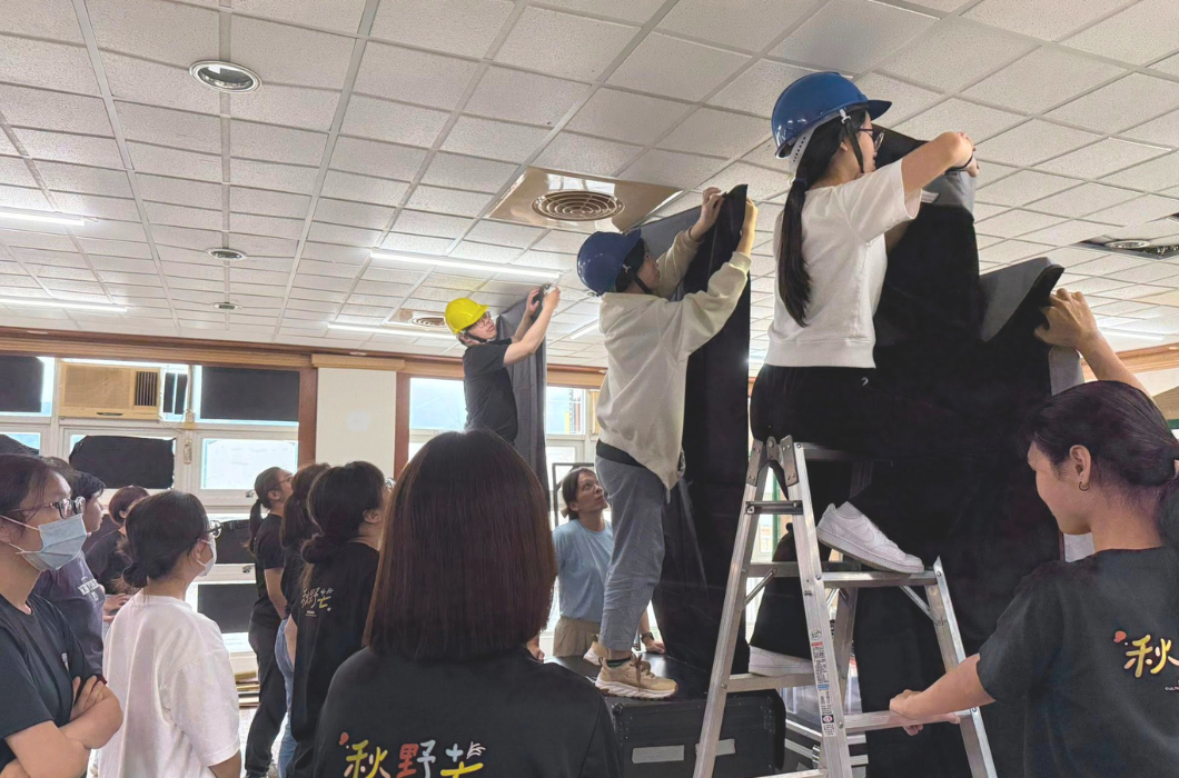 工作人員在室內架設黑布幕。幾位戴著安全帽的劇團工作人員站在梯子與道具箱上，合力將黑色大布幕固定在教室天花板架上，台下有多人圍觀協助。