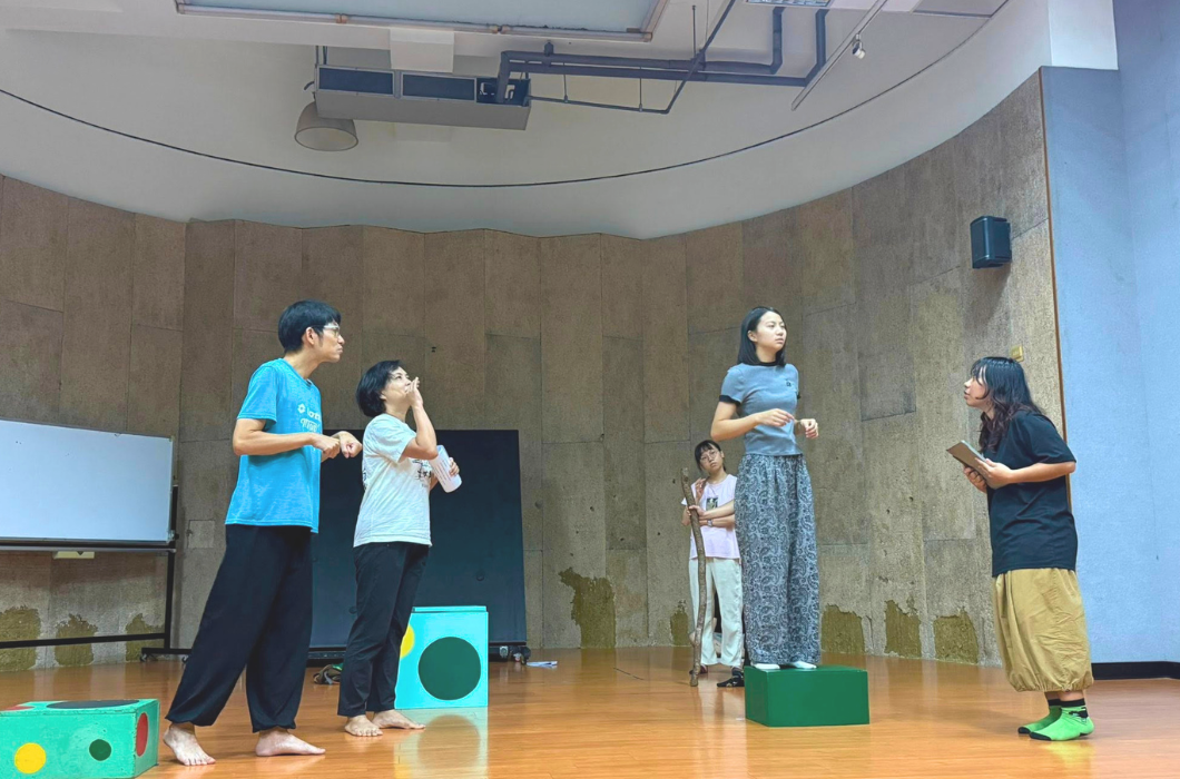舞台劇的排練現場。四位身穿便服的演員在排練室舞台上進行對手戲與走位調整，其中一位女性演員站在綠色道具方塊上，旁人正專注指導。