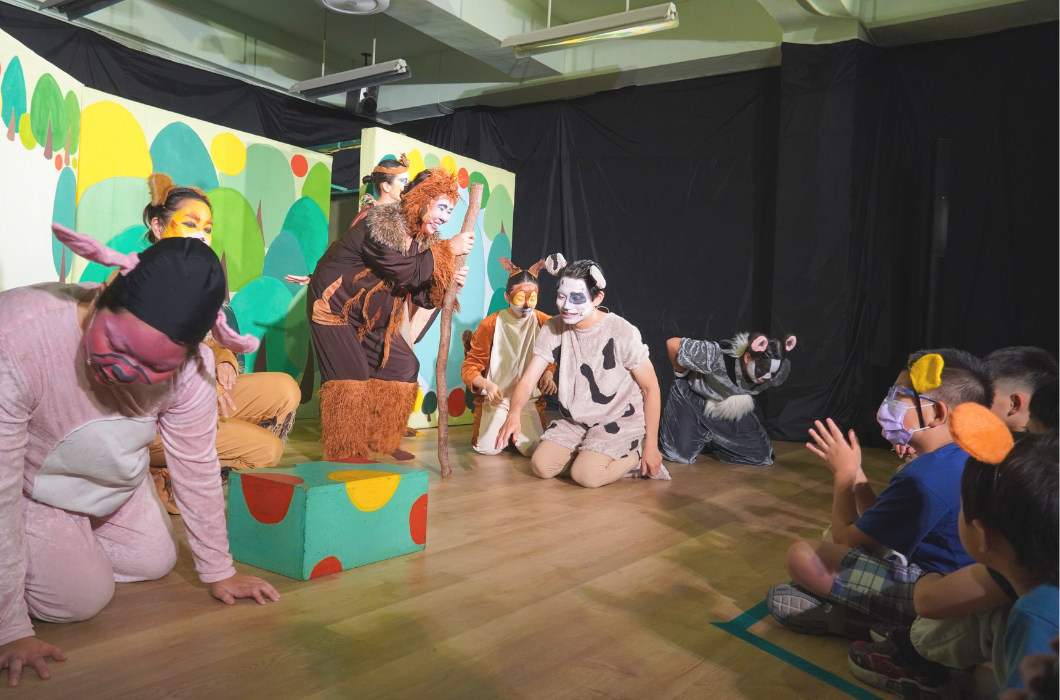 舞台劇正式演出側拍。舞台上多位穿著豬、狗、猴子等動物道具服的演員正圍在道具箱旁生動演出，右側的小朋友觀眾開心拍手互動。