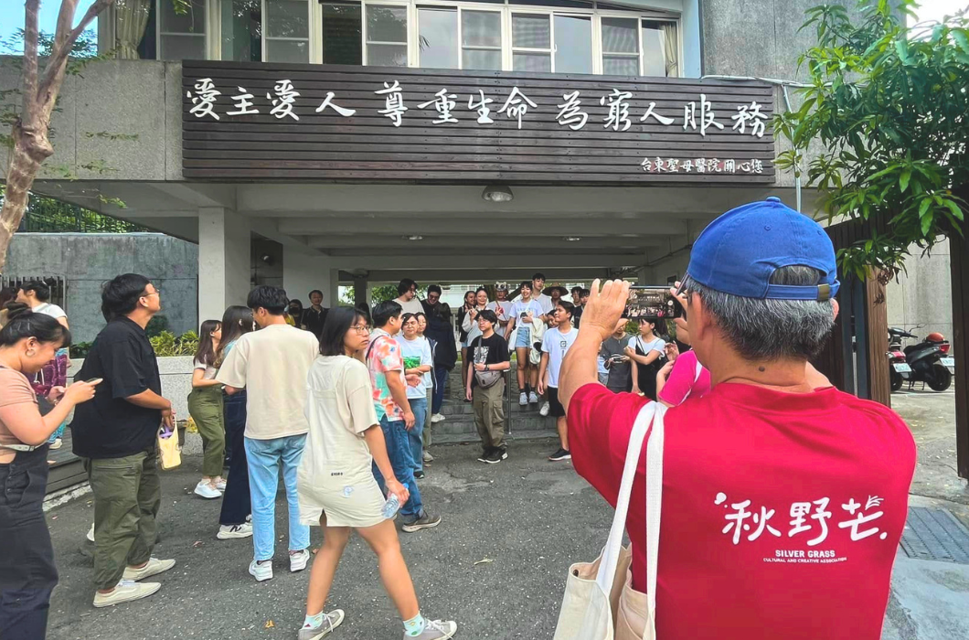 民眾聚集在台東聖母醫院的大門前，看板寫著「愛主愛人 尊重生命 為窮人服務」，前景有一名身穿「秋野芒」紅色上衣的男子正在用手機拍照。