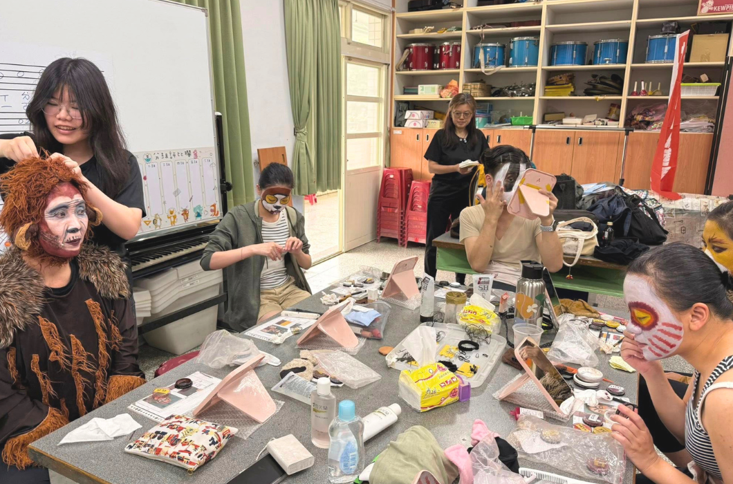 舞台劇後台，多位演員坐在桌前專心化妝，臉上塗著各式動物造型的彩繪臉譜。