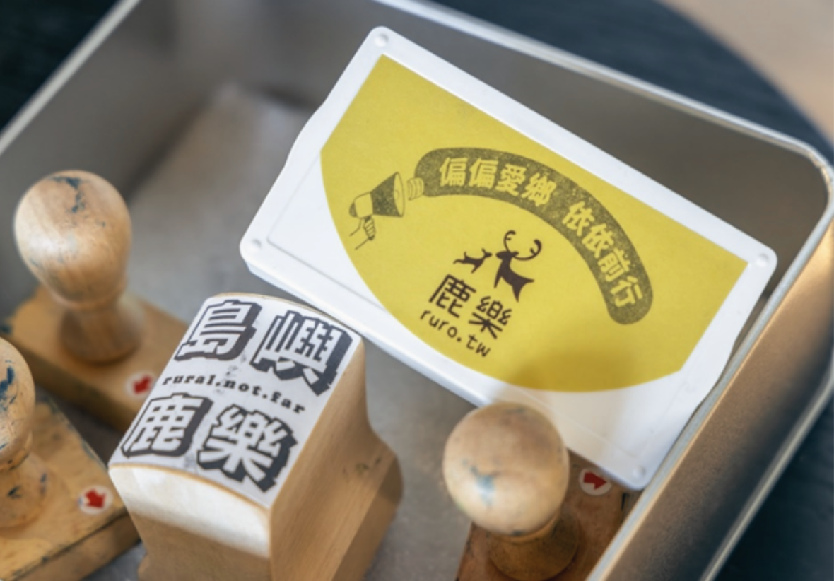 這張特寫照片展示了一個銀色金屬盒內的文創圖章組。主角是一個白框黃底的長方形貼紙，貼紙上印有手持擴音器的插圖，旁有文字「偏偏愛鄉 依依前行」。下方則印有大鹿與小鹿的剪影，以及文字「鹿樂 ruro.tw」。金屬盒內還有幾個木柄印章，其中一個木印章側面貼有白底黑字的貼紙，清楚寫著「島嶼鹿樂」及較小的英文字「rural.not.far」。照片前景左側還有另一個木印章，側面有紅色向下的箭頭貼紙。整體呈現一種溫暖的手作感與推廣鄉村教育或旅遊的意象。