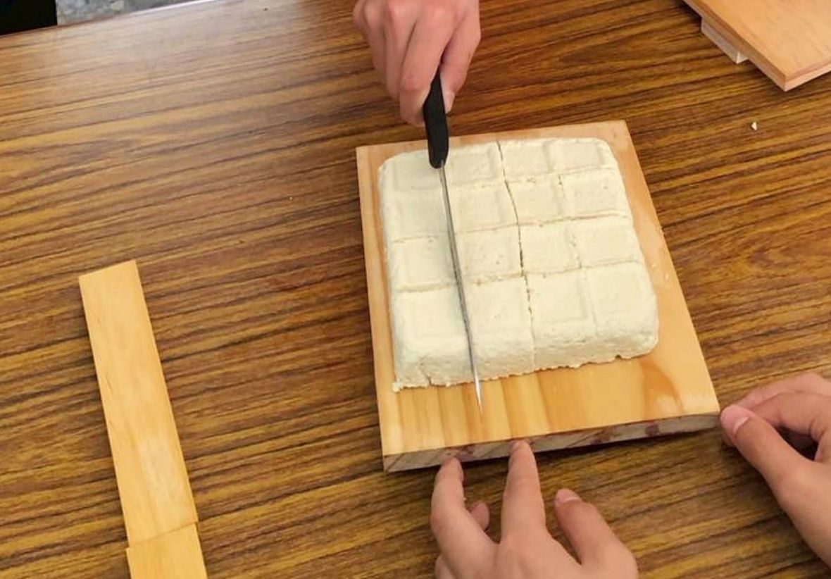 這是一張在木紋桌面上切豆腐的照片。畫面主體：一塊方形的白色板豆腐放置在淺色木砧板上。動作細節：上方的一隻手正手持刀子，將豆腐切成方格狀；下方則有另一雙手扶著砧板邊緣。背景環境：整體背景為深色木紋桌面，左側另有長條狀的木片。這張照片清晰呈現了手工切豆腐或製作豆腐的互動過程。