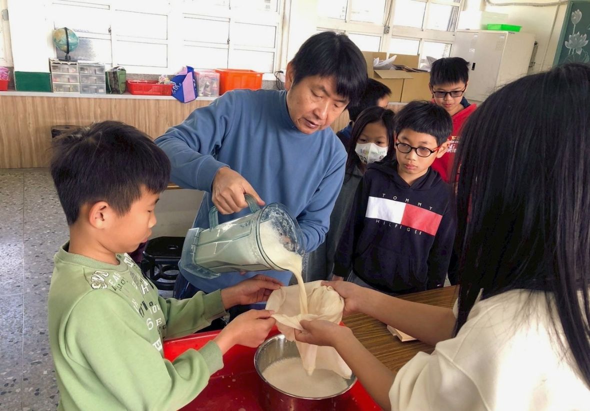 教室中，一名男老師正把果汁機裡的豆漿倒入濾布，兩名學生合力拿起濾布過濾豆漿，其餘三名學生在旁觀看。