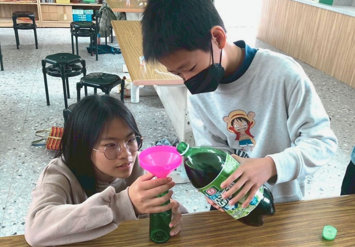 在教室內，一位戴口罩、穿著魯夫圖案衣服的男孩正將大瓶綠色飲料倒入女孩手持漏斗的玻璃瓶內，女孩戴著眼鏡專注看著瓶口。