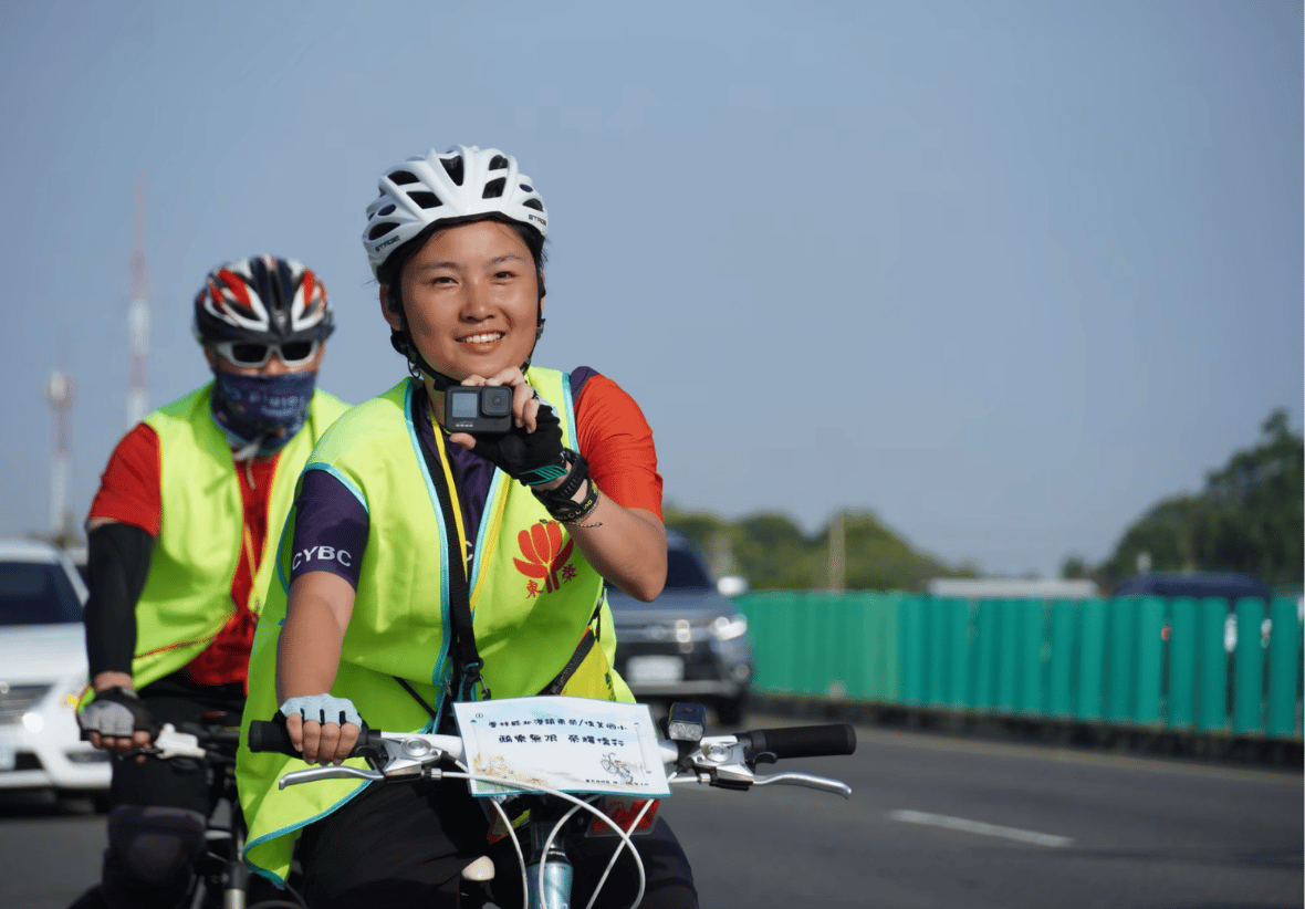 兩位身穿螢光綠反光背心、頭戴安全帽的單車騎士在寬闊的馬路上騎行。前方的女性騎士面帶微笑，右手正手持著一台小型運動攝影機記錄旅程；後方則有一位戴著墨鏡與黑紅安全帽的男性騎士緊隨其後，背景點綴著路旁護欄與遠方行駛的汽車。