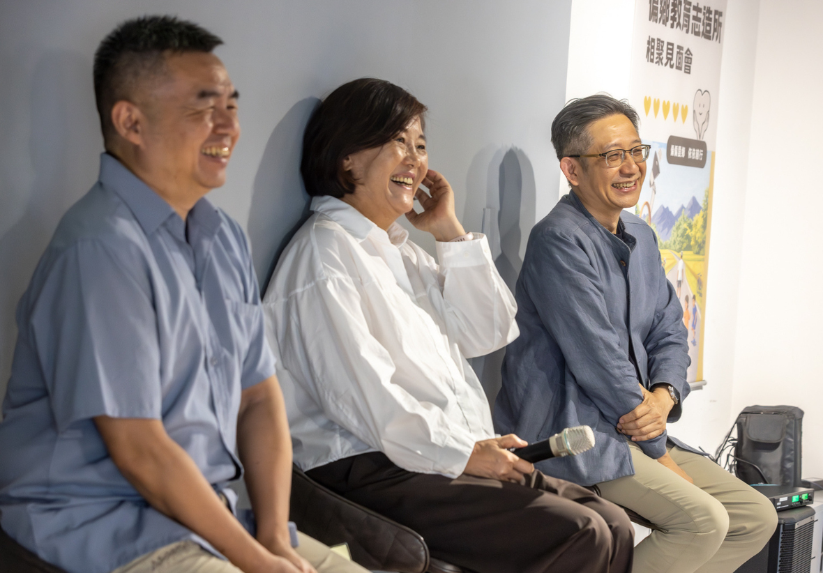 三位講者於活動現場並排而坐，氣氛愉快歡樂。左側男士穿藍色短袖襯衫燦笑；中間女士穿白色襯衫手持麥克風，笑著抬手摸頭髮；右側戴眼鏡男士穿藍色外套，雙手交疊膝上微笑。背景牆上掛有「偏鄉教育志造所 相聚見面會」的海報。