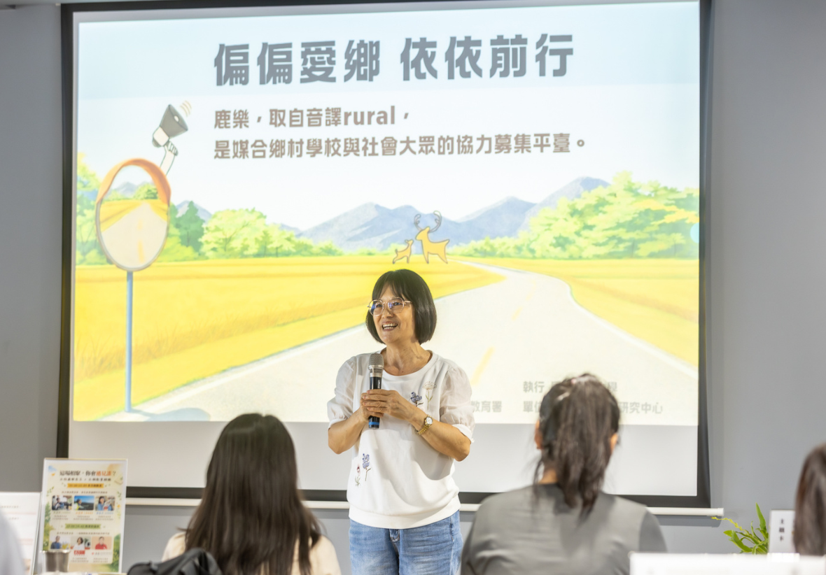 一名戴眼鏡、穿白色上衣與牛仔褲的女性講師手持麥克風在臺前演講，面對下方聽眾。背後大螢幕顯示簡報，標題為「偏偏愛鄉 依依前行」，內文寫著「鹿樂，取自音譯 rural，是媒合鄉村學校與社會大眾的協力募集平臺。」背景插畫為黃金稻田、道路與雙鹿圖樣。