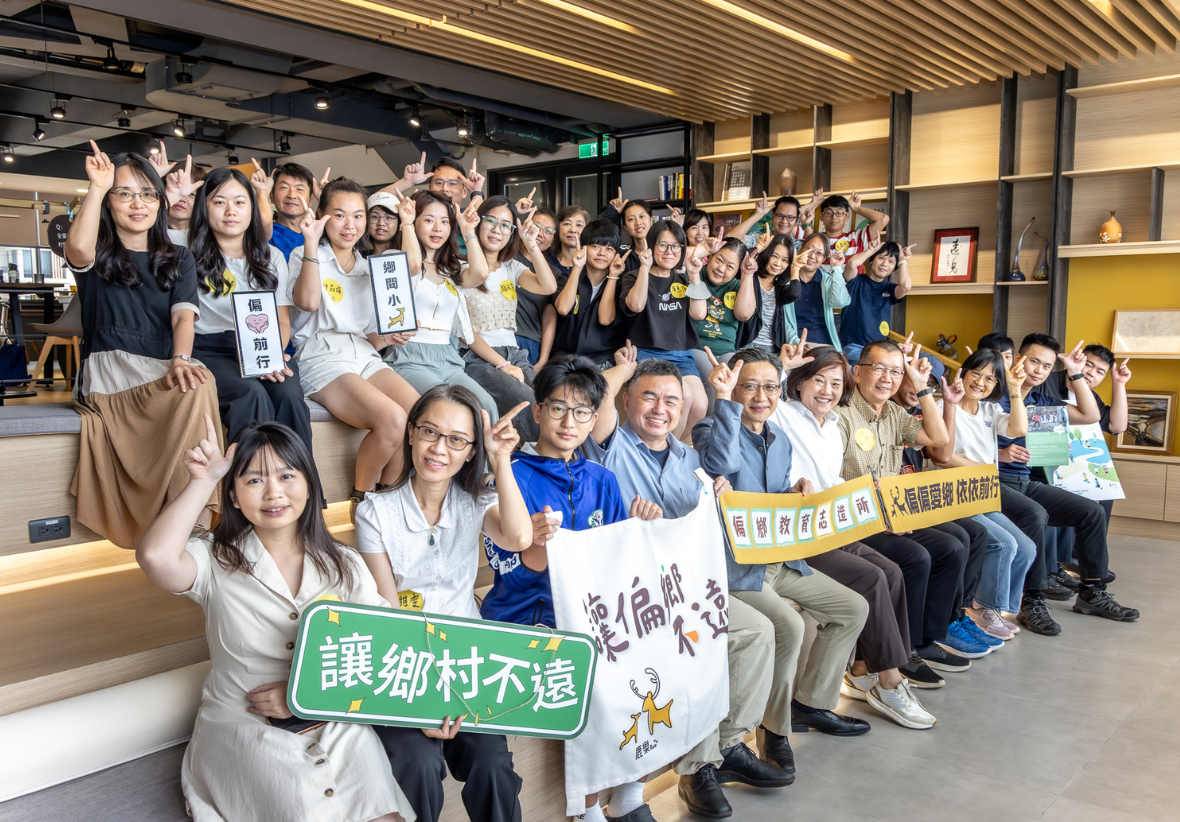 「偏鄉教育志造所」見面會的大合照。數十位參與者於階梯式座椅區歡樂合影，多數人頭頂比出鹿角手勢。前排人員展示「讓鄉村不遠」、「讓偏鄉不遠」、「偏鄉教育志造所」與「偏偏愛鄉 依依前行」等標語牌與布條，場面溫馨熱絡。