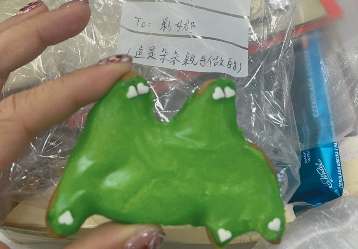 一隻手特寫持有綠色糖霜餅乾，餅乾上有白色心形圖案。背景可見透明塑膠袋和一張手寫字條，上寫著「To: 莉娜（這是朱朱親手做的）」。