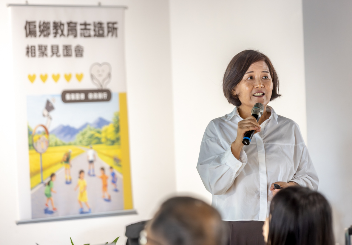 天下雜誌教育基金會執行長，手持麥克風在臺前演講，眼神專注。後方擺放「偏鄉教育志造所 相聚見面會」的海報，前景為聽眾背影。