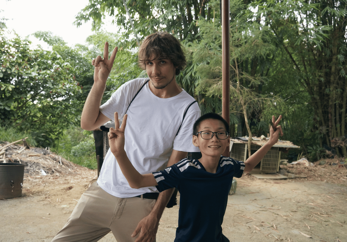 一位穿白色 T 恤的外國青年與一位戴眼鏡穿運動服的亞洲男孩，在竹林前開心合照。兩人皆面帶微笑，並雙手比出勝利手勢