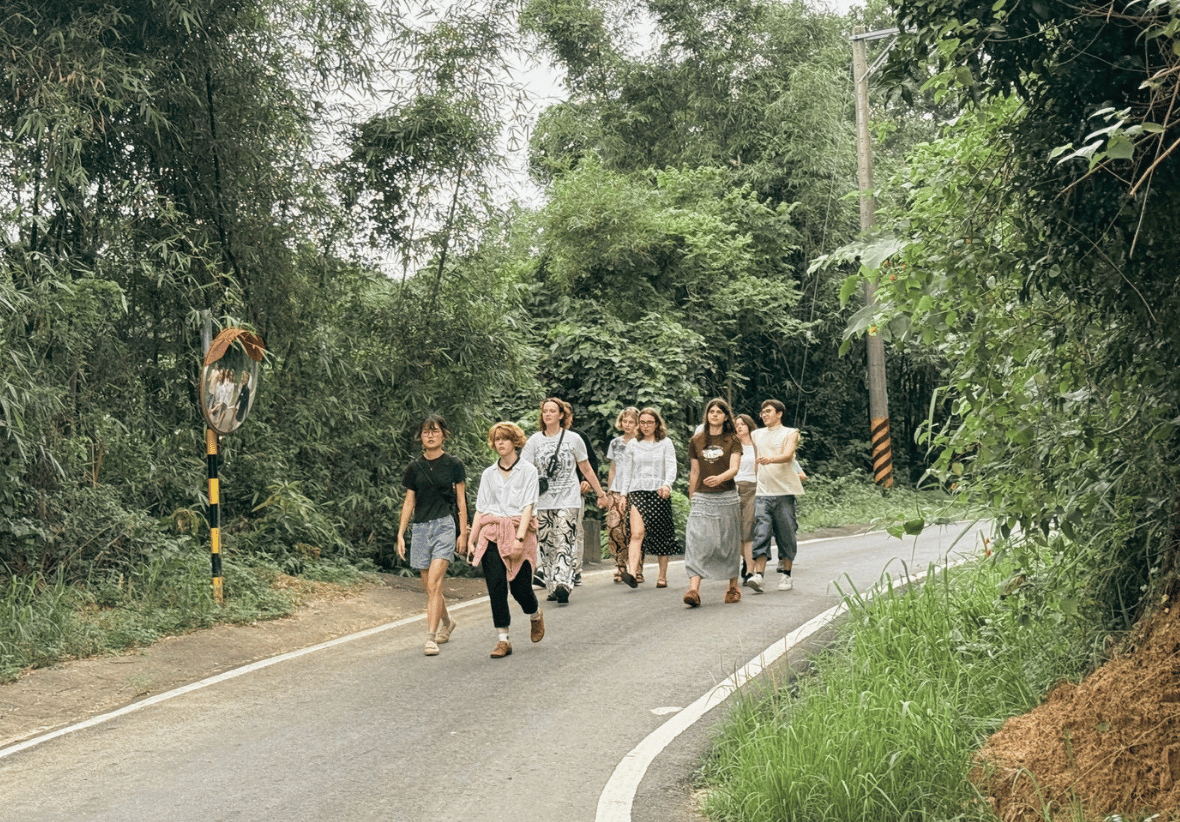 一群波蘭大學生沿著一條蜿蜒的鄉間柏油路行走。道路兩旁是茂密的竹林和綠色植被，路邊可見反光鏡。