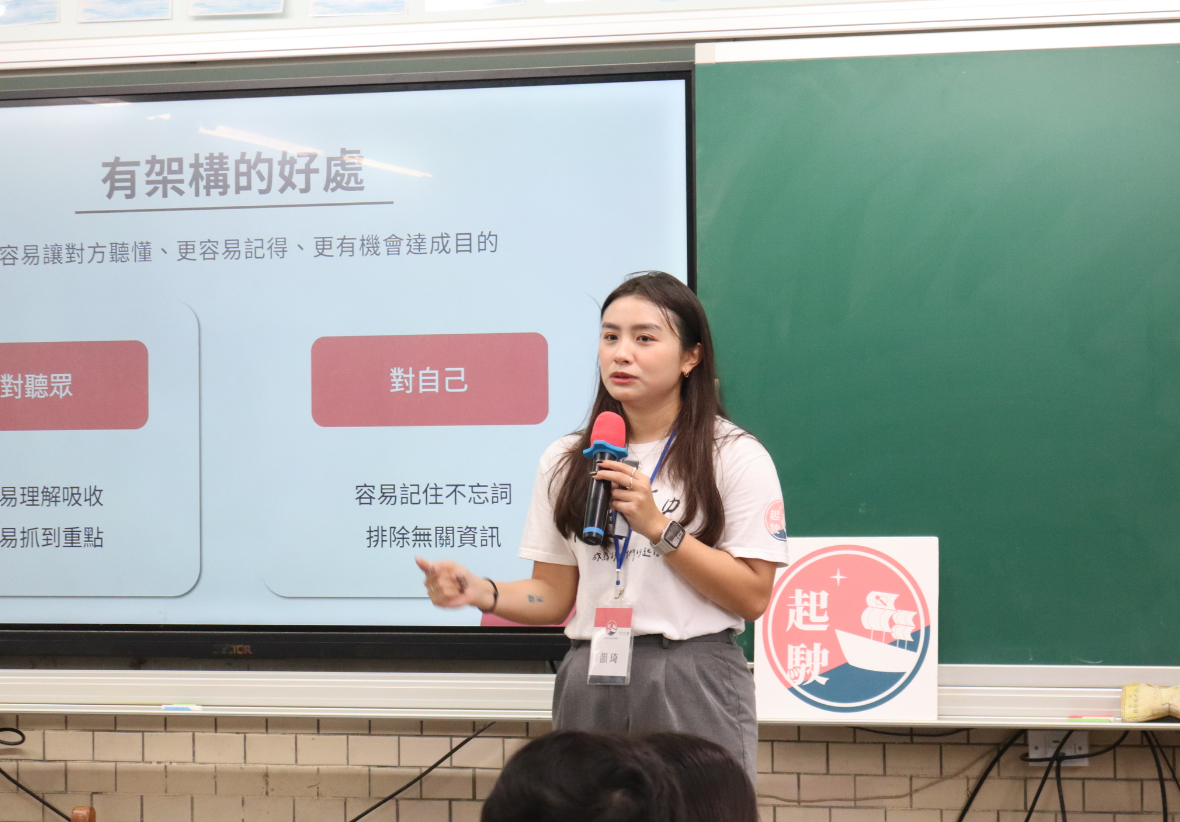一位年輕女性講師在教室黑板前手持麥克風進行授課，身後螢幕正顯示「有架構的好處」簡報頁面，簡報內容分為「對聽眾」與「對自己」兩大架構優點說明。