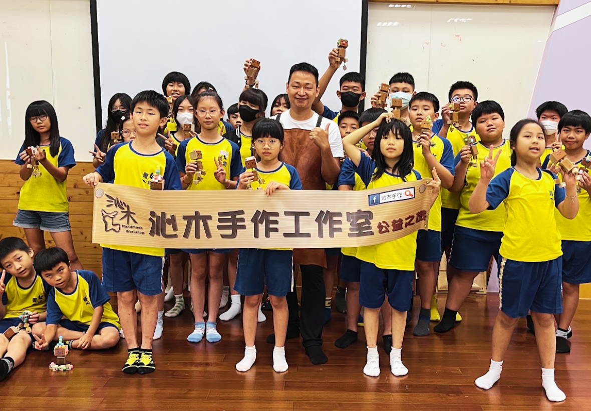 老師與一群身穿黃藍運動服的學生拉著「沁木手作工作室公益之旅」橫幅，開心地展示手作木偶並合影。
