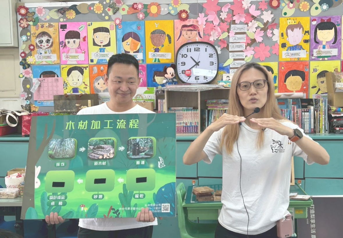 兩位講師在教室前進行解說，左側男講師展示「木材加工流程」的教學解說板，右側女講師配合手勢進行說明。