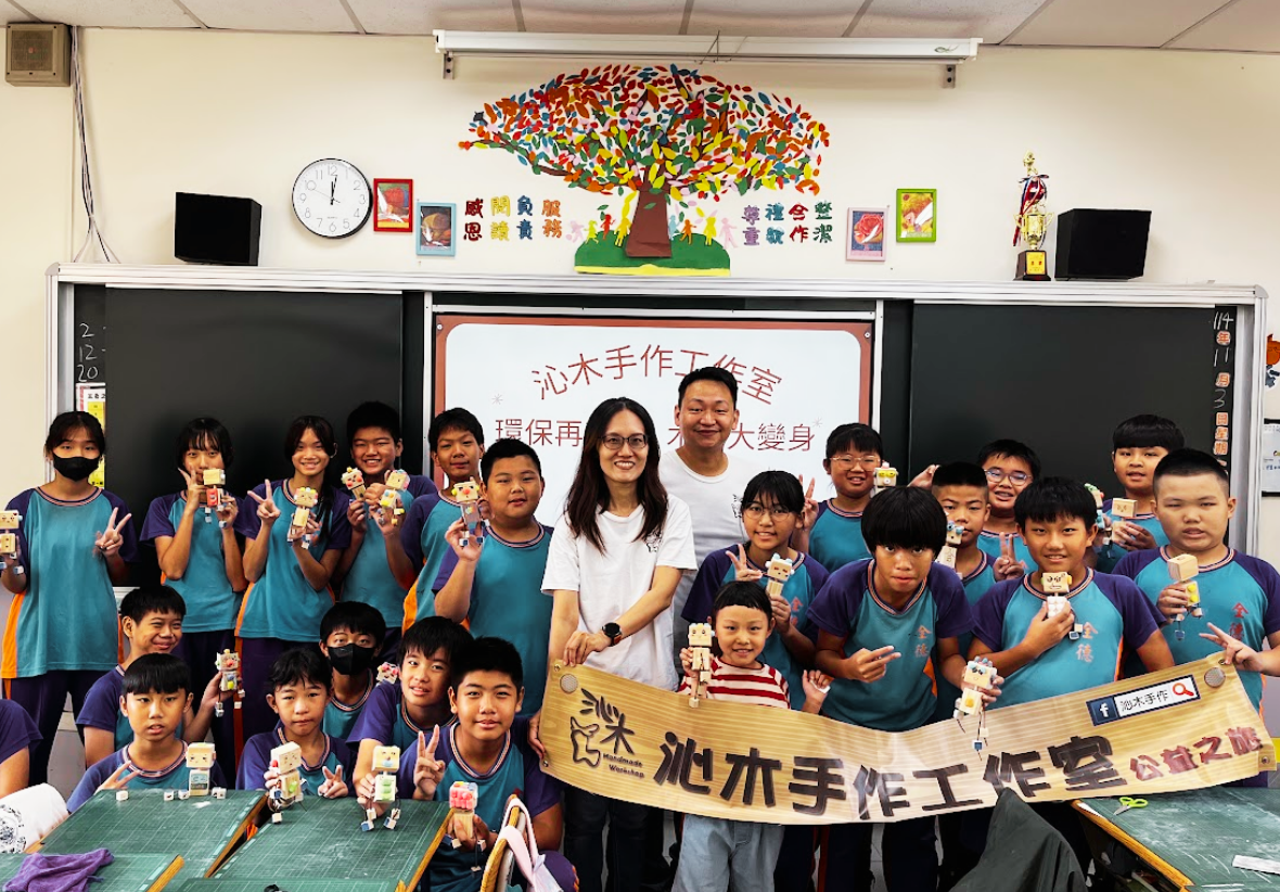 教室課堂大合照，兩位講師與全班學生一同拉著「沁木手作工作室公益之旅」橫幅，學生們開心地展示各自完成的木頭機器人作品。