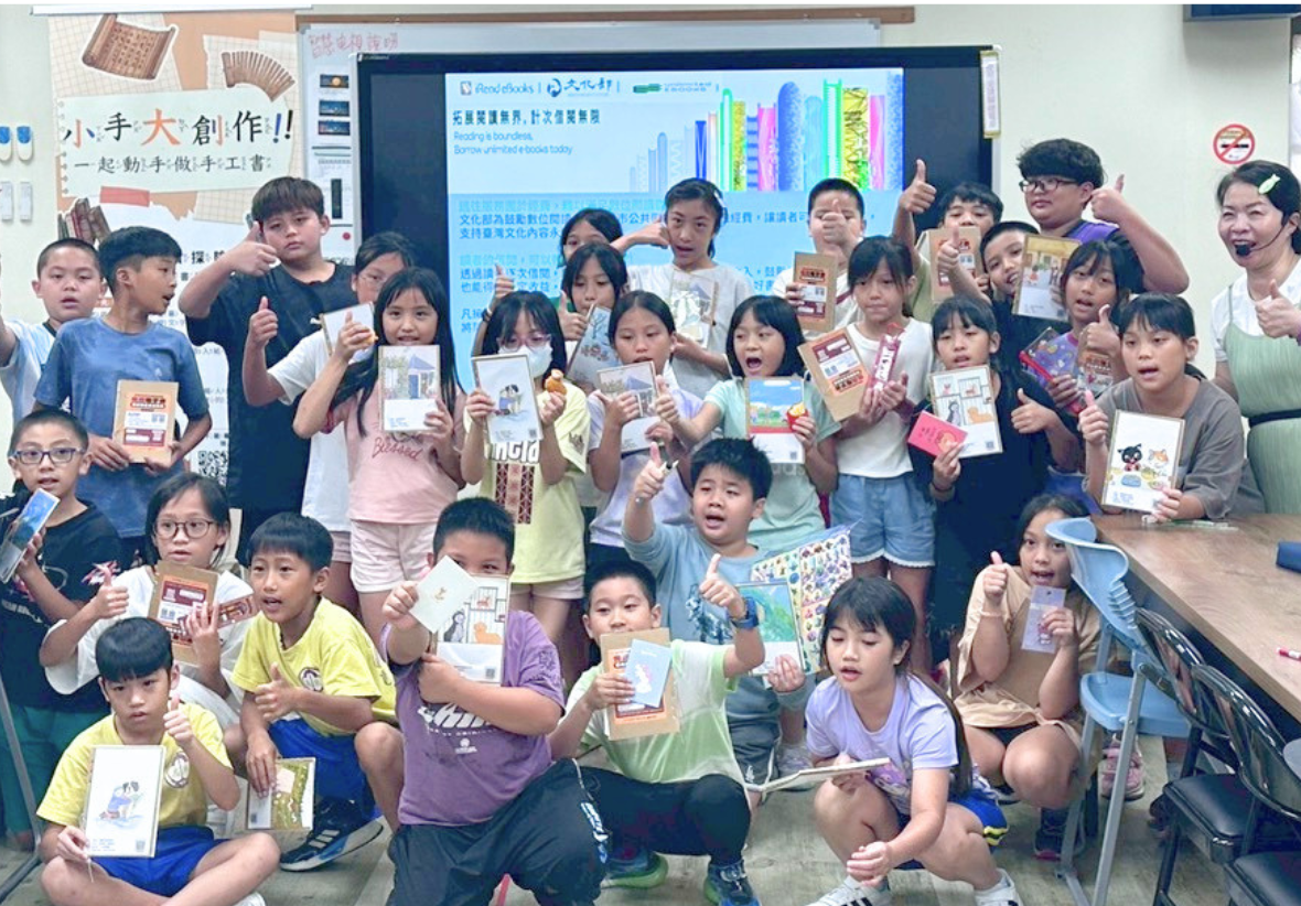 這是一張師生合照。教室內大約二十多位小學生和兩位女老師一起面向鏡頭。學生們坐在地板上，雙手大拇指比讚，並自豪地展示他們親手製作的小手工書。