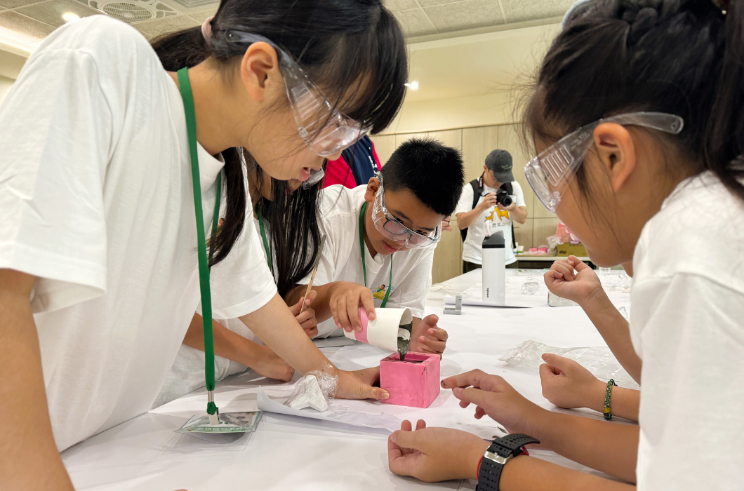 三名孩子戴著透明護目鏡，圍在桌邊分工合作，將混合材料倒入粉紅色模具，體驗低碳建材的製作過程。