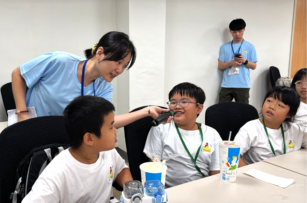女主持人彎下身將麥克風遞向戴眼鏡的男孩，鼓勵他分享想法；身旁兩名同伴轉頭聆聽，神情專注。