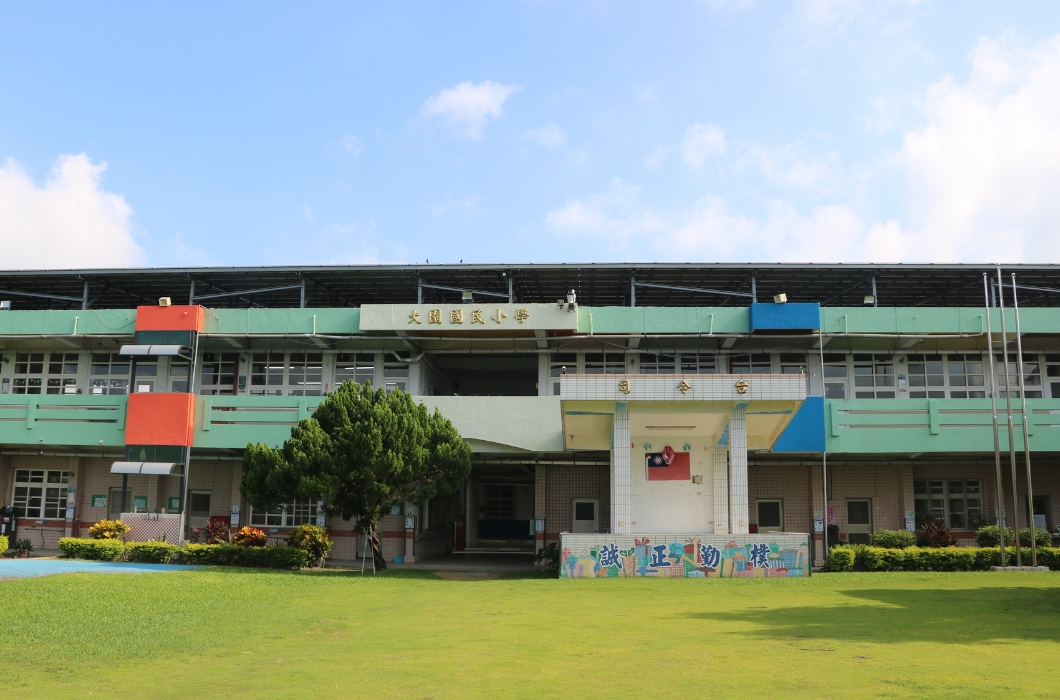 大園國民小學校舍外景，綠色與彩繪裝飾的建築前有草坪、司令台與大樹，天空晴朗。