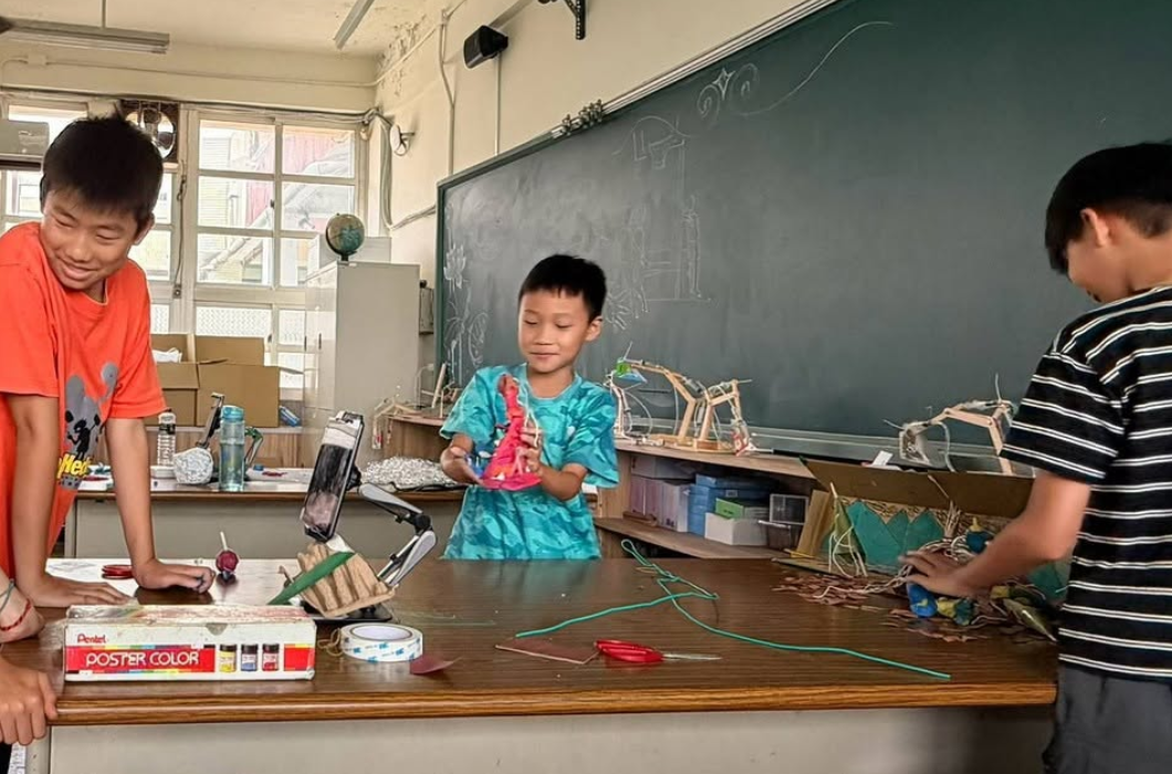 黑板前的課桌旁，幾位學童正在操作手機腳架，合力調整與拍攝桌上的立體黏土作品。