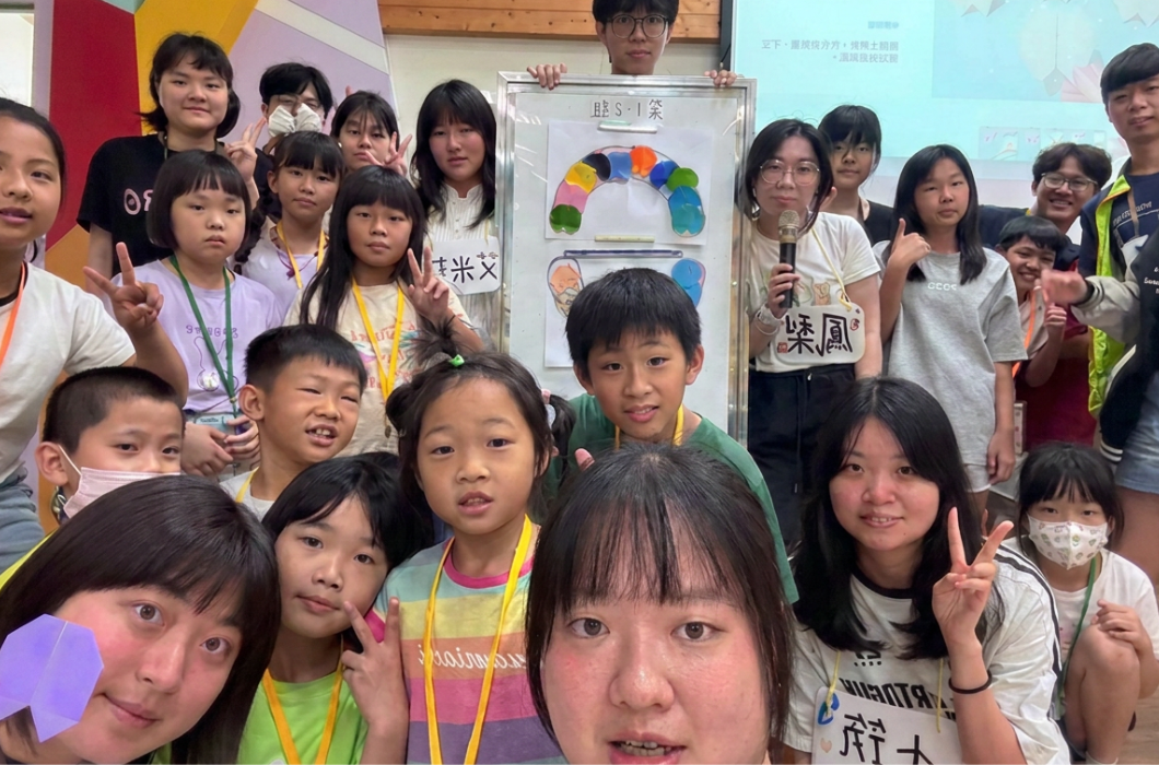 大小朋友圍繞著展示彩色圖畫的白板熱情合照，前方學童與後方隊員開心地面向鏡頭比出比手勢，氣氛活潑融洽。
