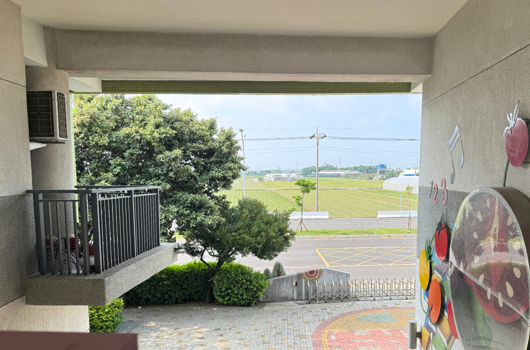 從校園二樓半戶外走廊陽台俯瞰，陽光灑在整齊的樹木、寬闊的街道與遠方一片翠綠的田園景緻，視野十分遼闊。