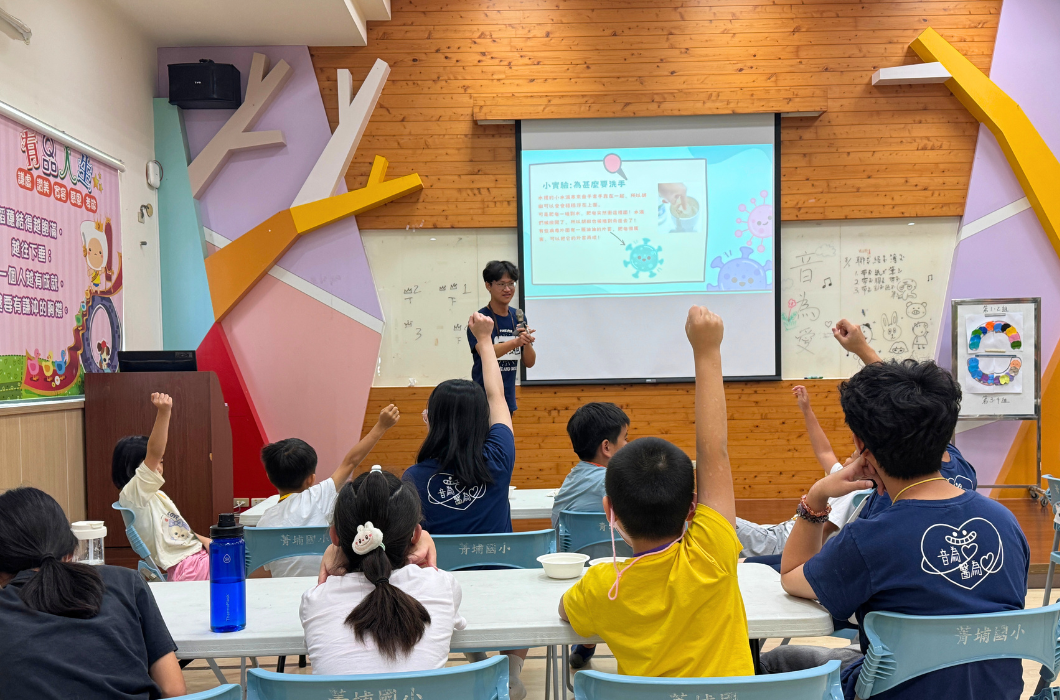 男講師在台上結合螢幕簡報進行洗手衛教課程，台下許多學生與隊員紛紛高舉手臂踴躍舉手發問，互動十分熱烈。
