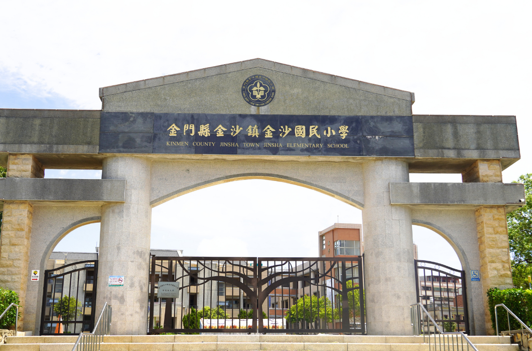 金門縣金沙鎮金沙國民小學的石柱校門，門額刻有校名，中央為鐵製大門與階梯，背景為晴朗的天空與校舍。