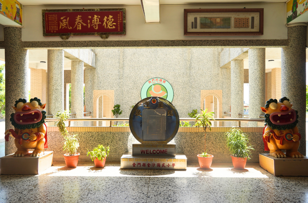金門縣金沙國民小學校園川堂，中央擺放迎賓標誌與盆栽，兩側設有可愛造型的風獅爺雕像，上方懸掛「德溥春風」匾額。