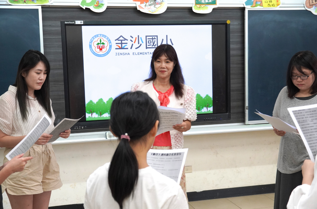教室電子白板前，老師與幾位學生圍成一圈，每人手中皆拿著稿紙，正在進行朗讀或朗誦練習。