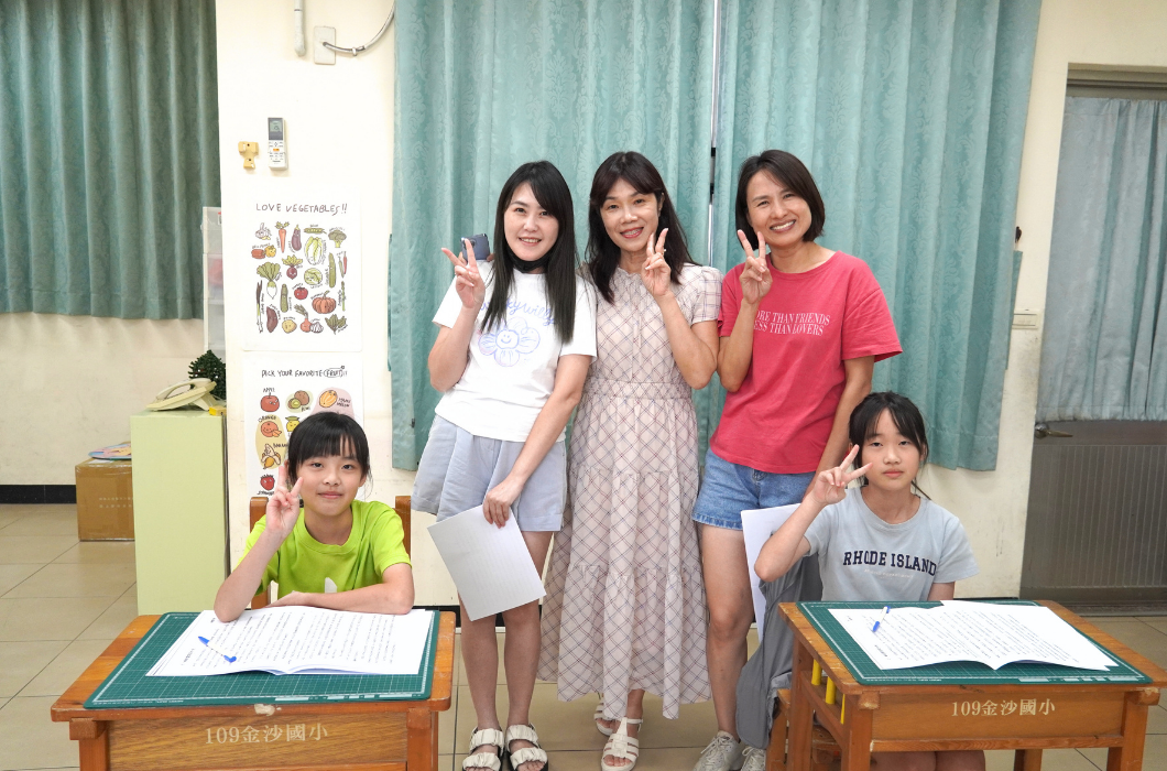 三位女性與兩位坐在課桌前的學童在教室內合照，大家對著鏡頭比出 V 字手勢，展現歡樂氛圍。
