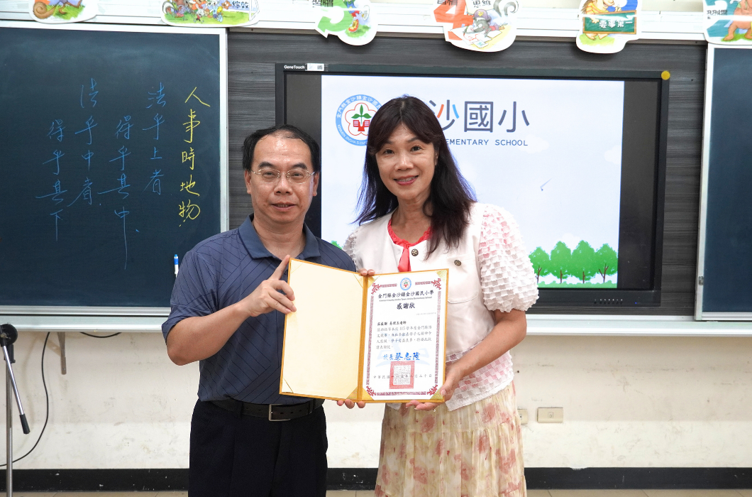 教室電子白板前，一位穿著深色 polo 衫的男性與老師共同高舉金門縣金沙國民小學頒發的感謝狀合影。