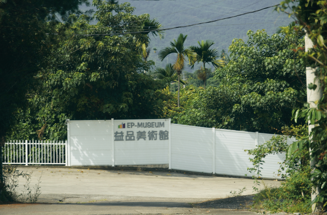 柏油路底，一堵白色格柵圍牆上嵌有『EP-MUSEUM 益品美術館』字樣及標誌。背景是綠樹與遠山