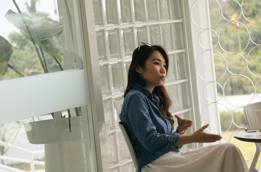 一名穿牛仔外套的女子，戴筱薇總經理坐在白色椅子上，在玻璃磚與鋼琴烤漆圓環裝飾的窗前，手勢豐富地說話
