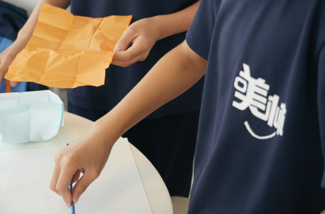 身穿印有「美林」字深藍 T 恤的人正拿筆在紙上繪畫，旁邊的人雙手展示著展開的橘色摺皺紙。