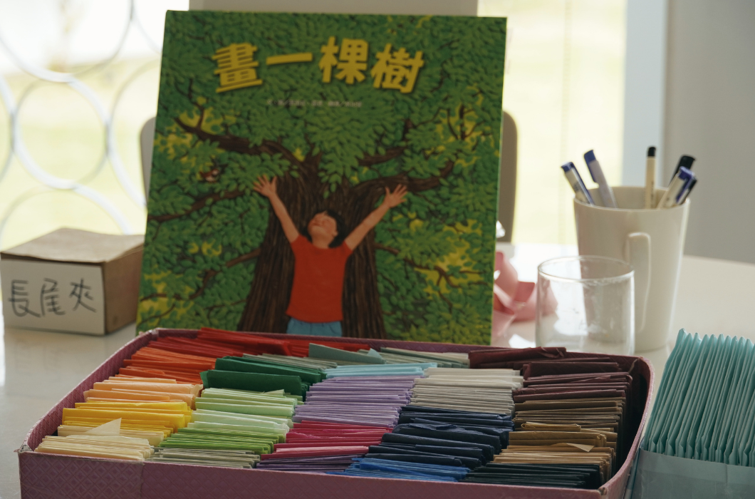 桌上擺著封面寫有《畫一棵樹》的繪本，前方盒子分類擺滿紅、黃、綠、紫等各色整齊的彩色紙片。