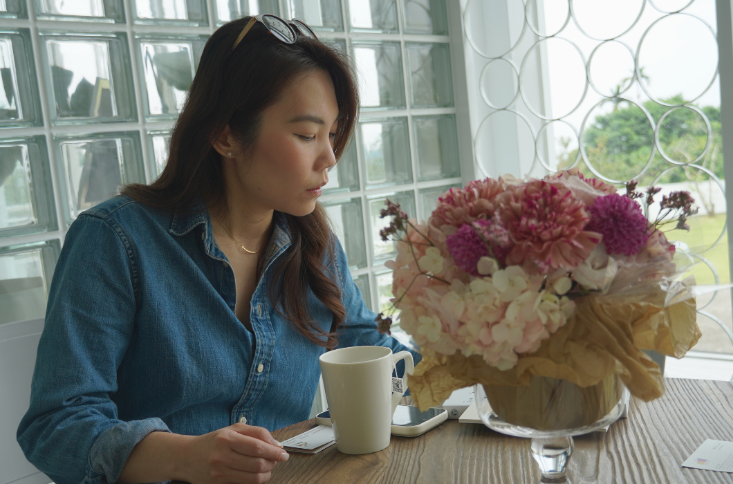 身穿牛仔襯衫的女子坐在桌前低頭專注，桌上擺放著一杯茶水與一盆粉紫色系的大束鮮花。