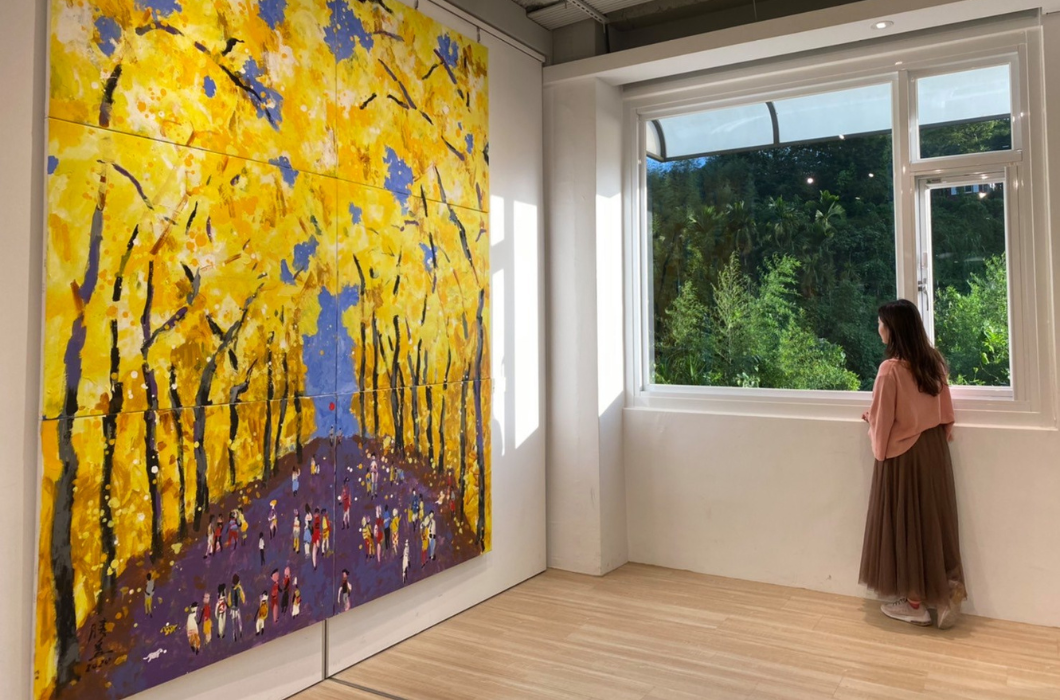 展廳牆上掛著大型黃色銀杏樹畫作，一名女子站在右側大開窗前，望向窗外的綠意山景。