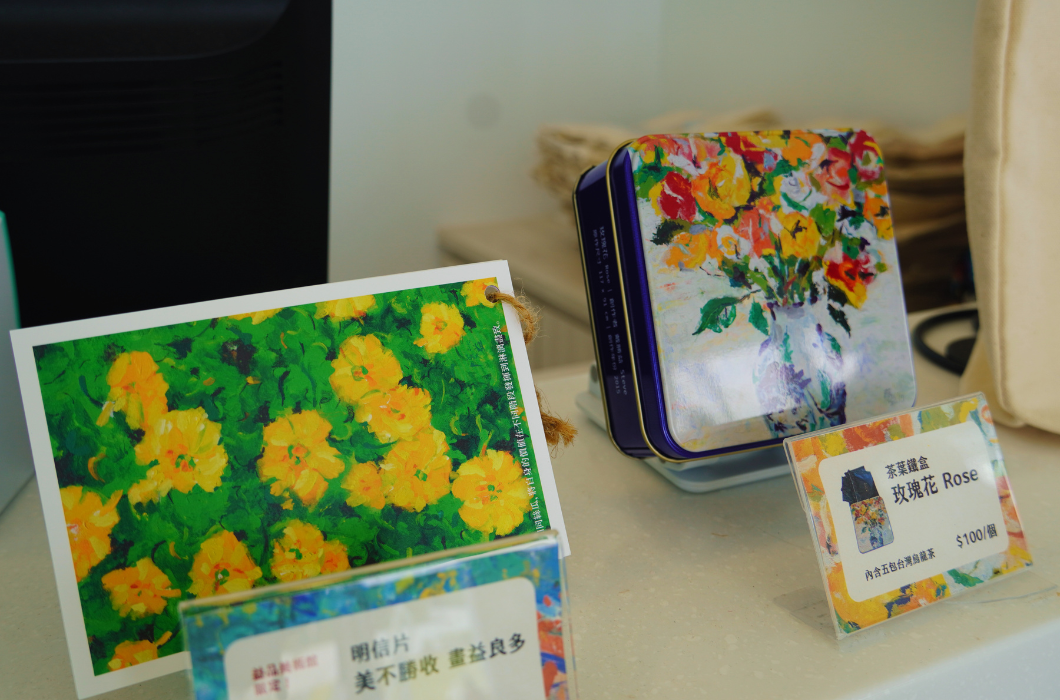 桌上展示周邊商品：前方為黃花畫作明信片，右側為印有花卉油畫圖案的藍色茶葉鐵盒與價格標示牌。