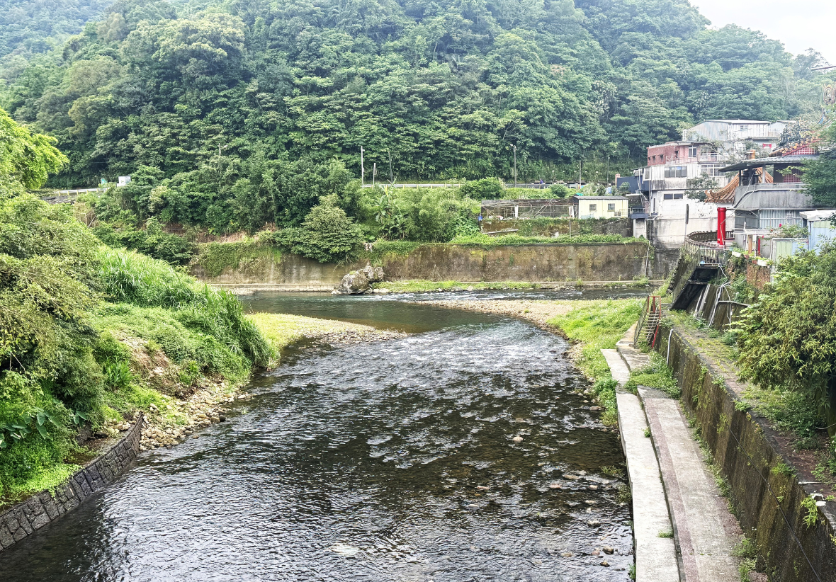 溪流在綠意茂密的山腳下交會，水面泛起細小波紋，岸邊露出碎石淺灘，右側沿岸可見民宅、廟宇與步道。