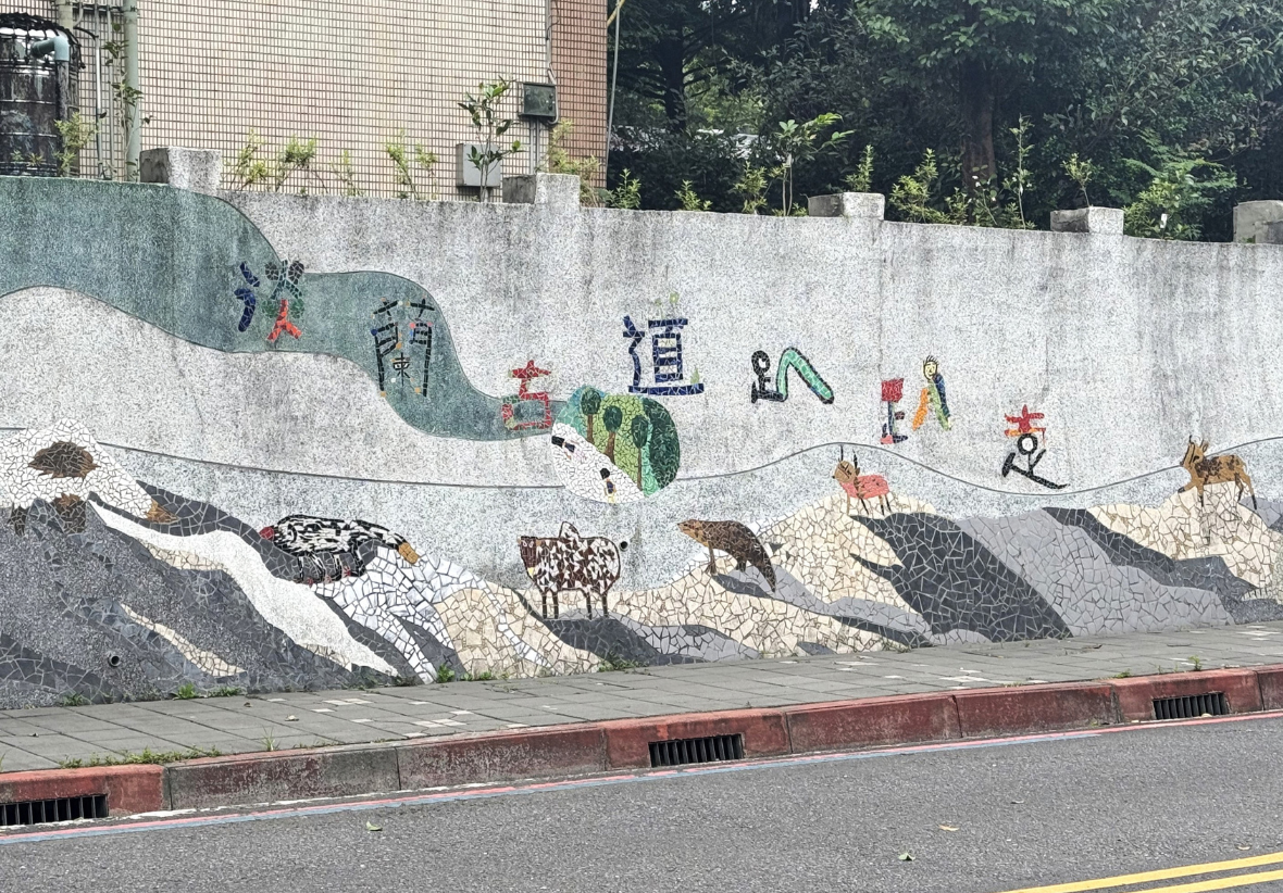 路旁長牆以馬賽克拼貼蜿蜒山形、人物與多種動物，上方搭配彩色中文字，牆前是人行道，後方可見建築與樹林。