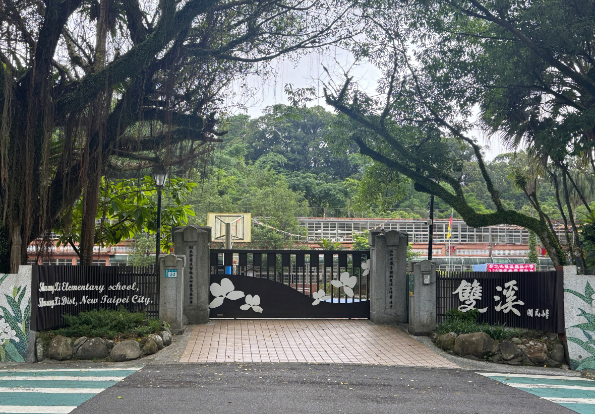 雙溪國小校門掩映在大樹枝葉下，黑色鐵門飾有白色花朵，右側標示「雙溪」，門後可見籃球架與校舍。