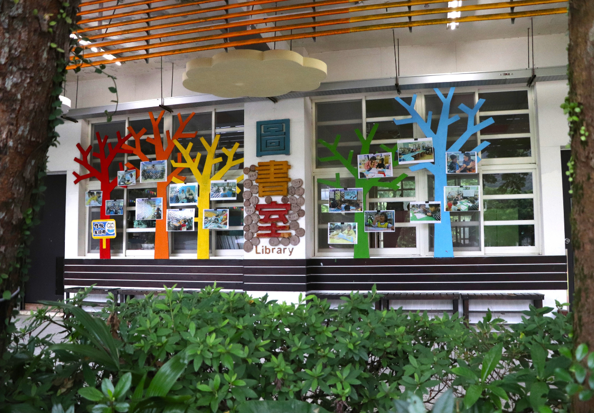 圖書室外牆裝飾著紅、橙、黃、綠、藍色樹木，枝幹間展示學生活動照片，中央標示「圖書室」，下方設有長椅。