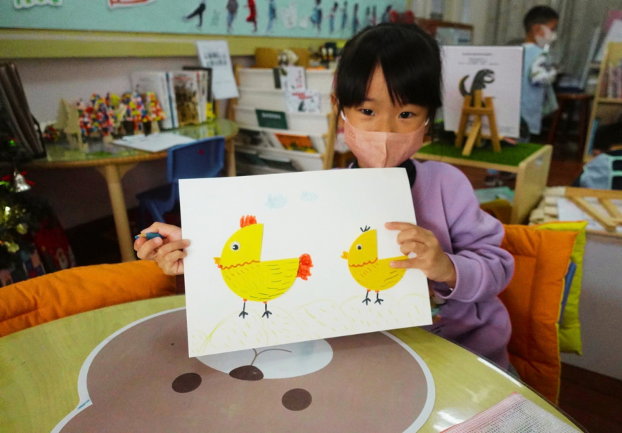 戴粉紅色口罩、穿紫色上衣的小女孩坐在桌前，雙手展示自己的畫作，紙上貼繪著一大一小兩隻黃色小雞，背景為擺放圖書與教具的教室。